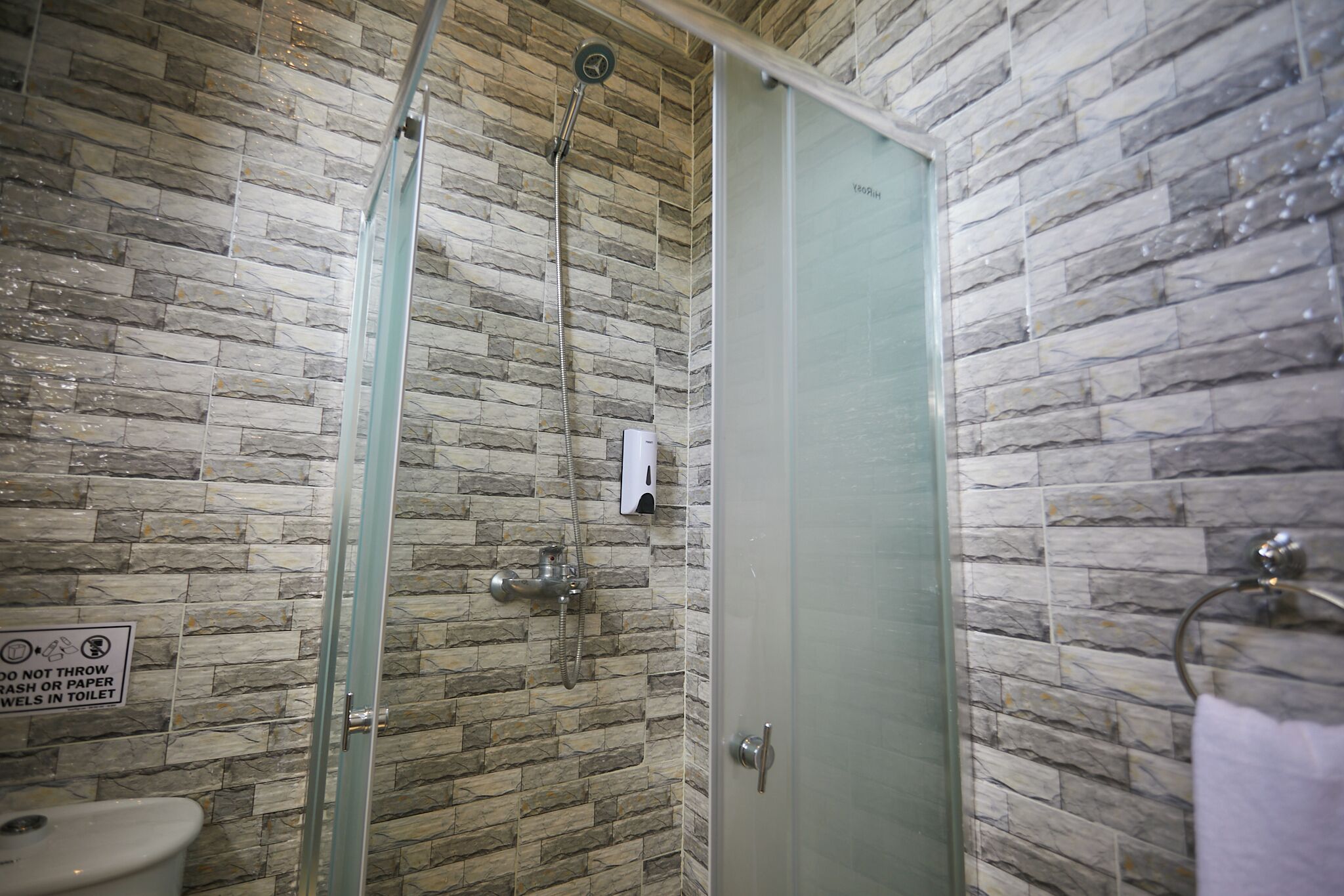 A bathroom with a stone wall, a glass shower enclosure, a toilet, and a hand soap dispenser.