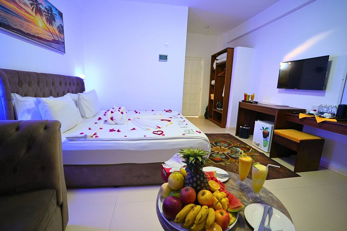 Habitació d'hotel amb un llit, una taula de menjador amb fruita i begudes, una televisió i un armari.
