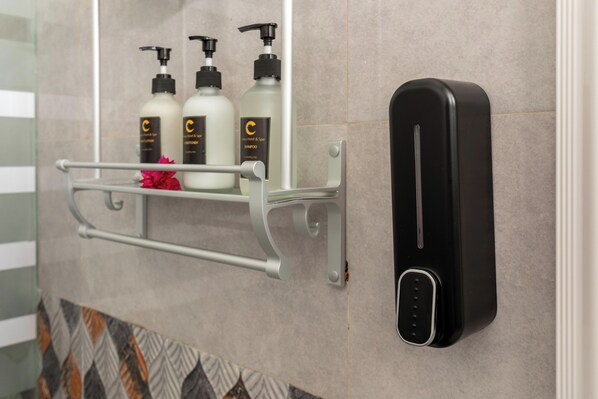 A bathroom with a hand sanitizer dispenser mounted on the wall, a metal shelf holding three bottles of shampoo, and a patterned tile backsplash.