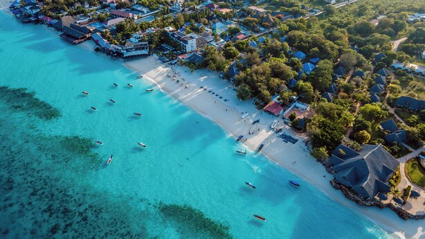 Vue aérienne d’une station balnéaire avec une plage, des eaux turquoise et limpides, et plusieurs bateaux.