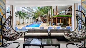 Un espace aménagé au bord de la piscine, avec des chaises longues, une table en verre et une vue sur un immeuble doté de balcons et de palmiers.