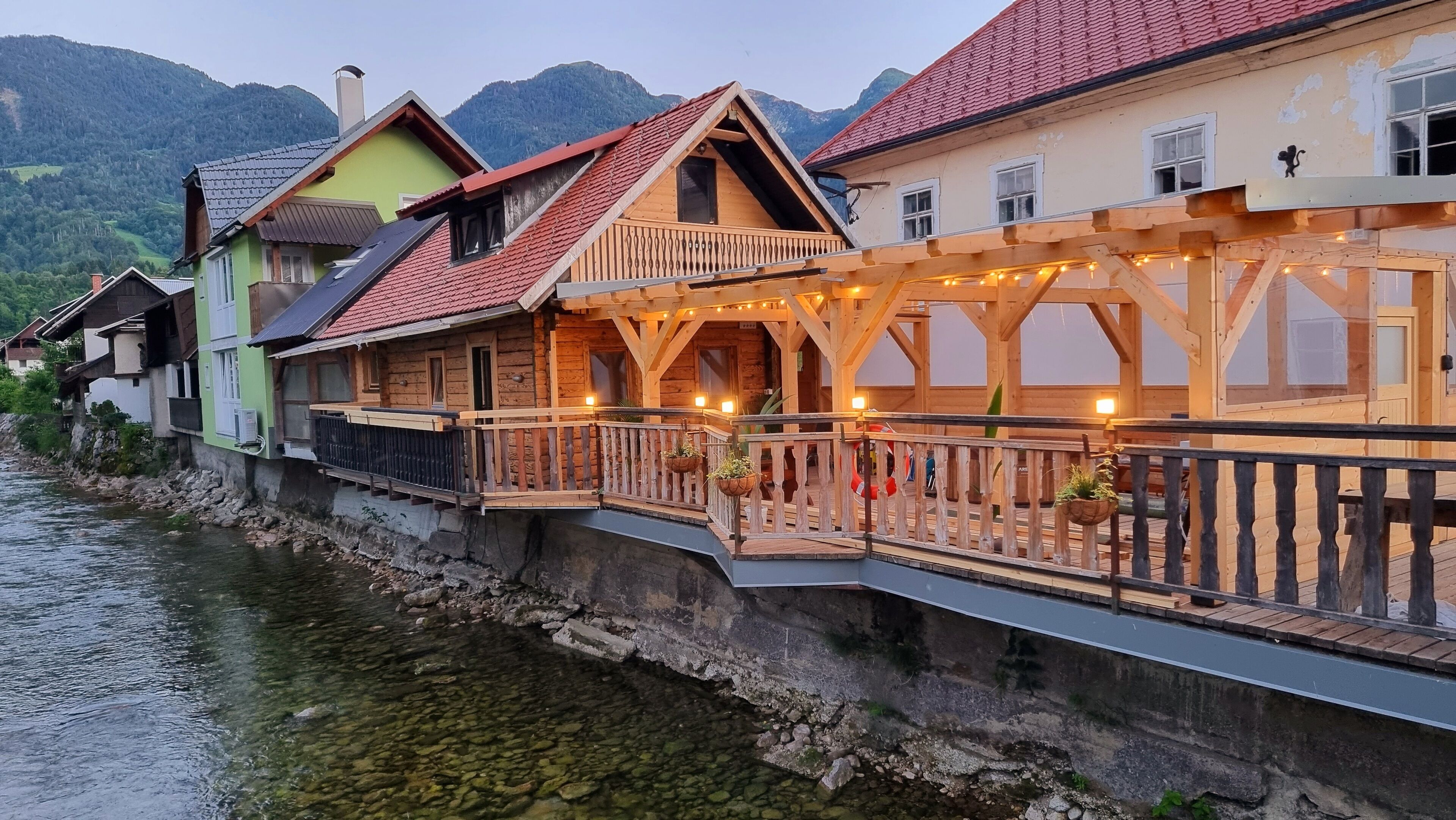 Fileira de casas de madeira com varandas voltadas para um rio, com montanhas ao fundo.