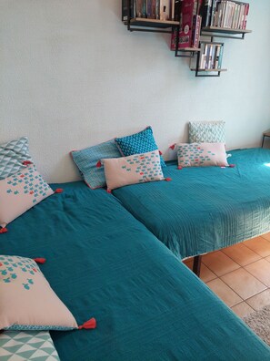 Una habitación con dos camas, cada una con ropa de cama color turquesa y almohadas estampadas. Un estante con libros está fijado a la pared.