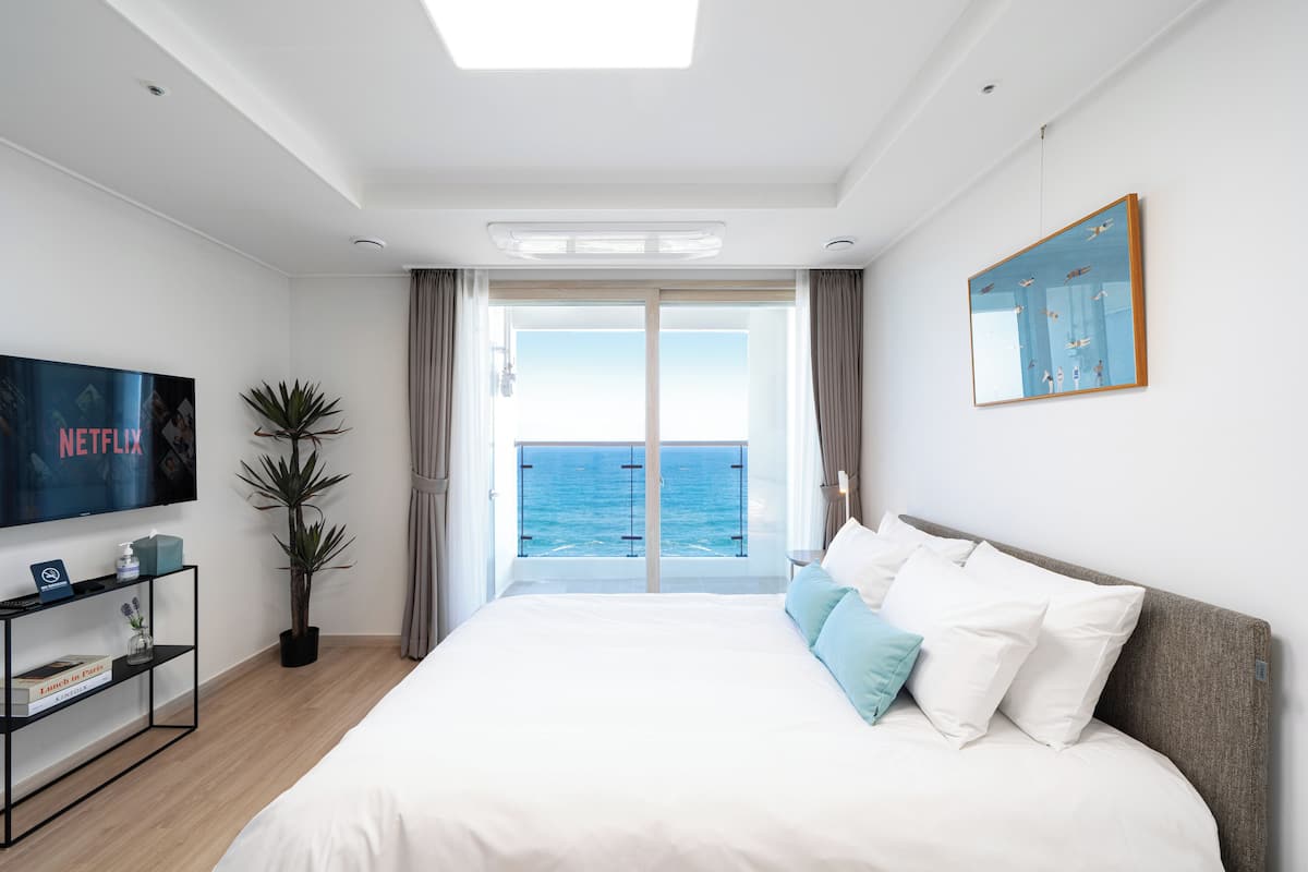 Quarto de hotel moderno com uma cama grande, televisão, planta em vaso e vista para o mar por uma porta de correr.