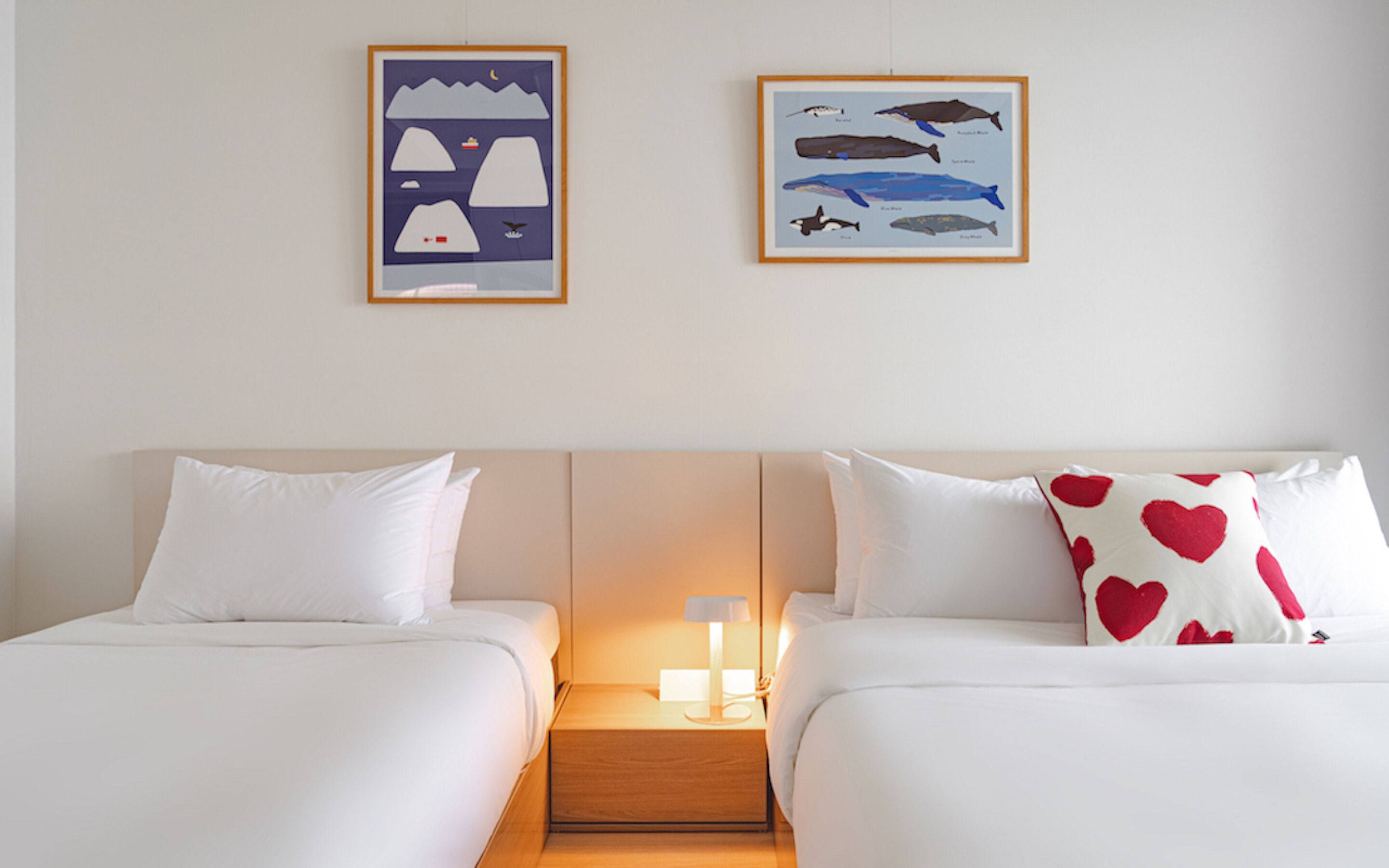 A hotel room with two beds, white bedding, a nightstand with a lamp, and two framed pictures on the wall.