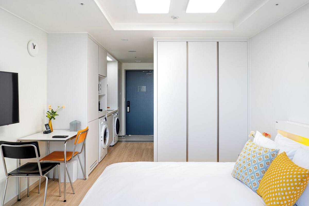 Quarto de hotel moderno com cama, escrivaninha, cadeira, máquina de lavar e porta azul.