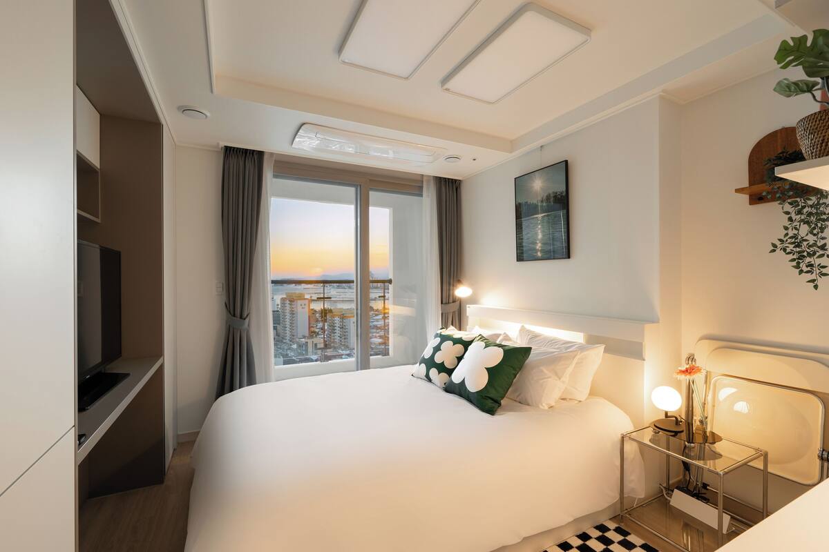 Quarto de hotel com uma cama grande, televisão, escrivaninha com luminária e vista para a cidade ao pôr do sol.
