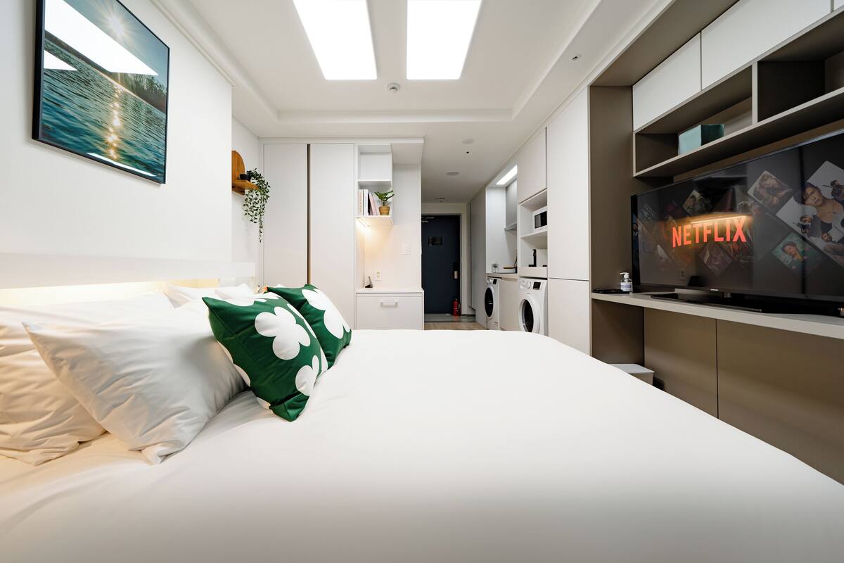 Quarto de hotel moderno com cama, televisão exibindo o logotipo da Netflix, área de cozinha e banheiro.
