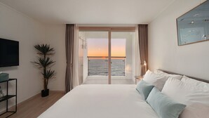 Een hotelkamer met een groot bed, een televisie, een potplant en uitzicht op zee bij zonsondergang.
