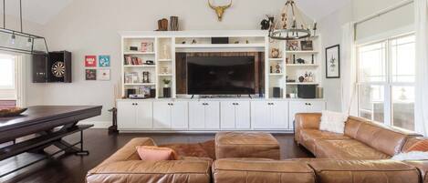 A spacious living room with a large leather sofa, a dining table, and a TV mounted on a white cabinet.