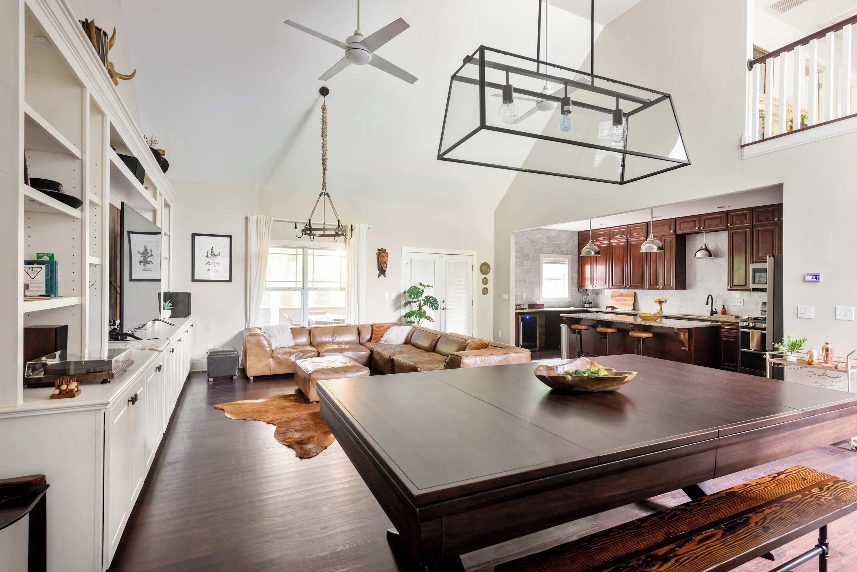 A spacious living area with a large wooden dining table, a leather sofa, and a kitchen with dark wooden cabinets and pendant lighting.