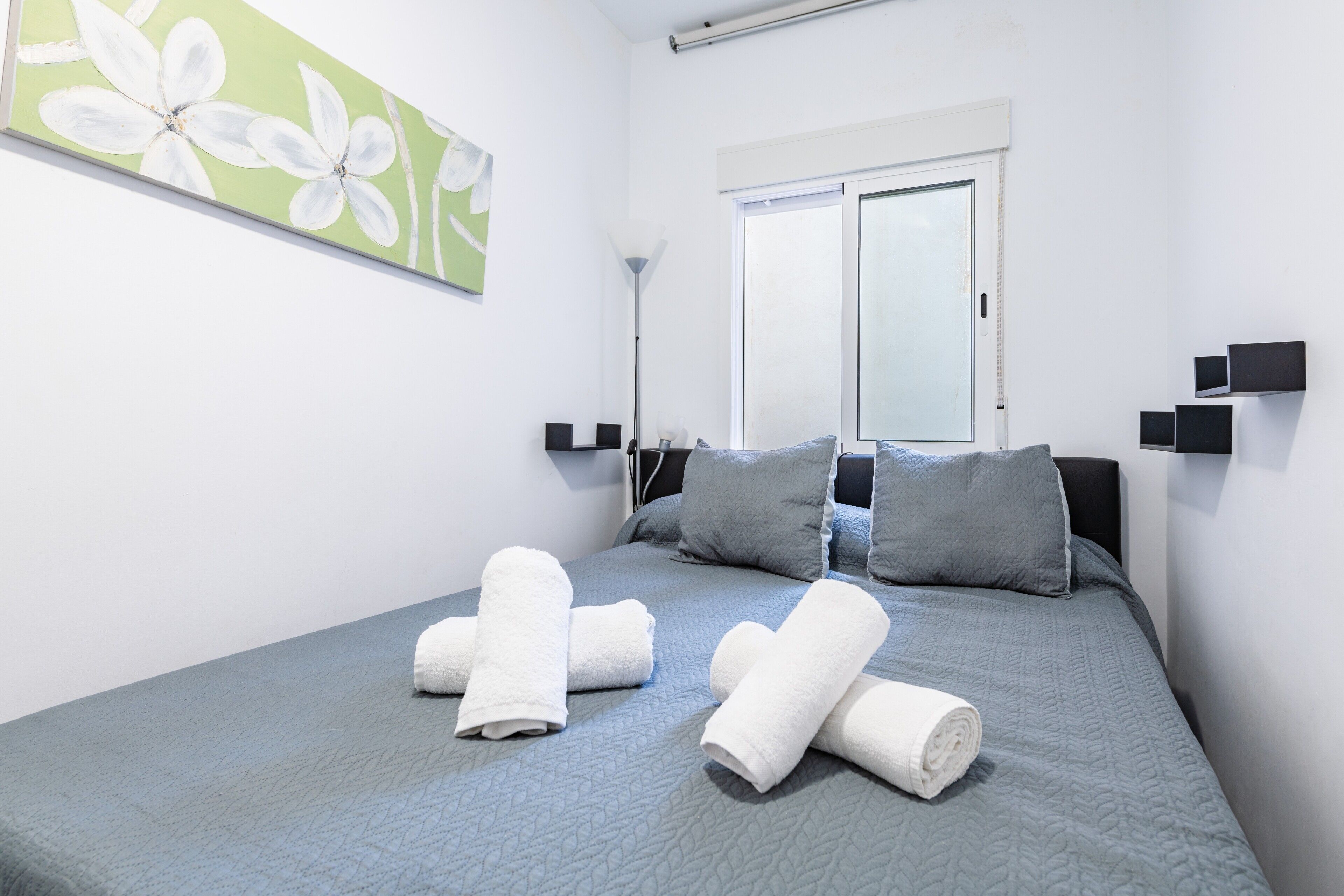 Una cama bien hecha con un edredón azul, una pared verde con un cuadro floral y una ventana con marco blanco.