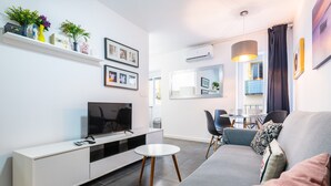 Ein modernes Wohnzimmer mit einer Couch, Fernseher, einer weißen Konsole und einem Essbereich im Hintergrund.