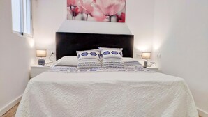 Una cama bien tendida con una colcha blanca y almohadas estampadas, flanqueada por dos lámparas de noche, en una habitación con suelo de madera y una gran obra de arte floral sobre la cama.
