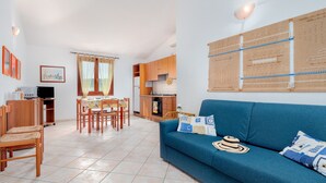 A living room with a sofa, a dining table with chairs, a kitchen area, and a television.