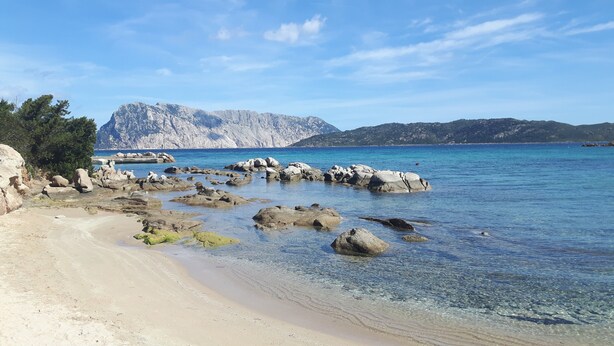 A beach with clear water, rocks, and mountains in the background.