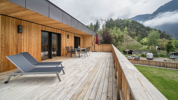 Una terraza de madera con una silla de descanso, una mesa con sillas y vistas a un paisaje montañoso.