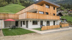 Una casa moderna de dos pisos de madera, con balcón, un porche cubierto con mobiliario exterior y vista a una ladera verde.