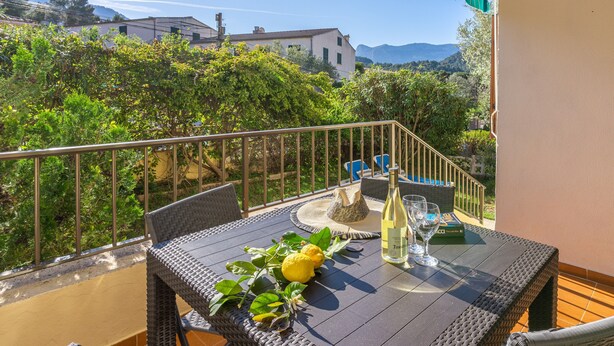 A balcony with wicker furniture, a table with a bottle and glasses, and a view of greenery and buildings. - Holiday Home "Yolima" near the Beach with Wi-Fi, Air Conditioning and Garden (Sóller)