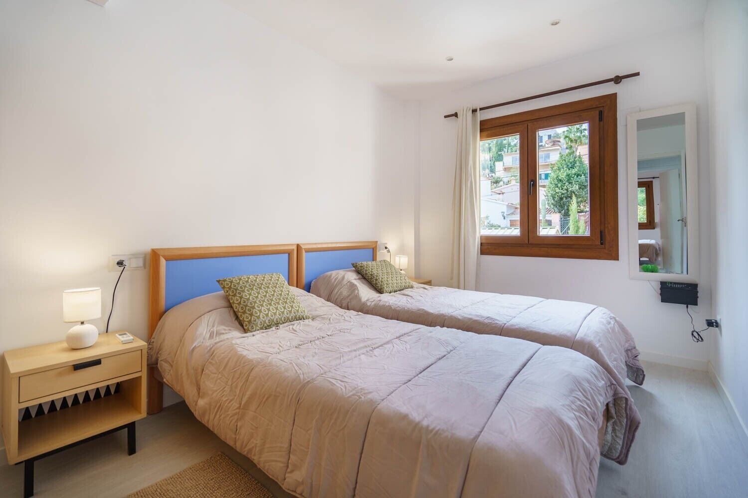 A bedroom with two beds, a nightstand with a lamp, a window with a view, and a mirror on the wall.