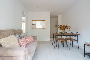 A modern living room with a sofa, a dining table with chairs, a mirror on the wall, and a vase with flowers.