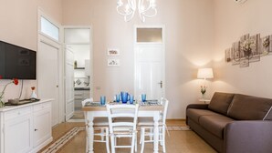 Una sala de estar con un sofá marrón, una mesa de comedor con sillas, un televisor y una araña.