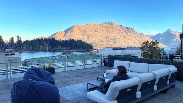 A rooftop terrace with a sofa and chairs, overlooking a lake and mountains.
