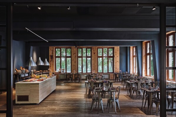 A spacious dining area with wooden tables and chairs, large windows, and a central buffet with various food items.