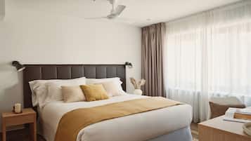 Une chambre d’hôtel moderne dotée d’un grand lit, d’un ventilateur de plafond, d’une table de chevet avec une lampe et offrant une vue sur l’extérieur à travers des rideaux légers.