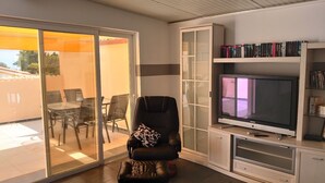 Una sala de estar con una puerta corrediza de vidrio que da a un patio exterior, un sillón de cuero negro, un televisor sobre un soporte de madera y una estantería con libros.