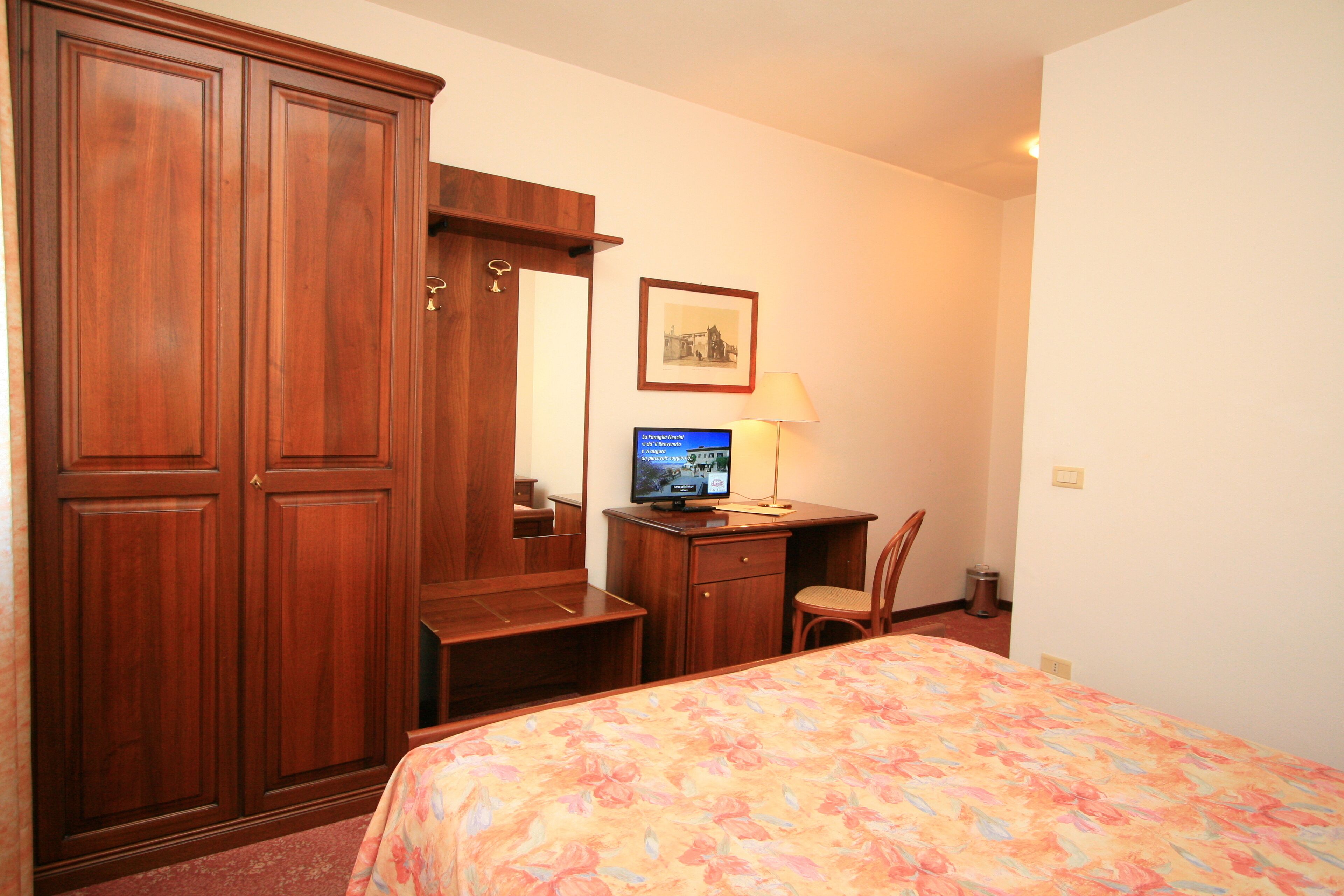 A hotel room with a bed, a desk with a computer, a chair, a wardrobe, and a framed picture on the wall.