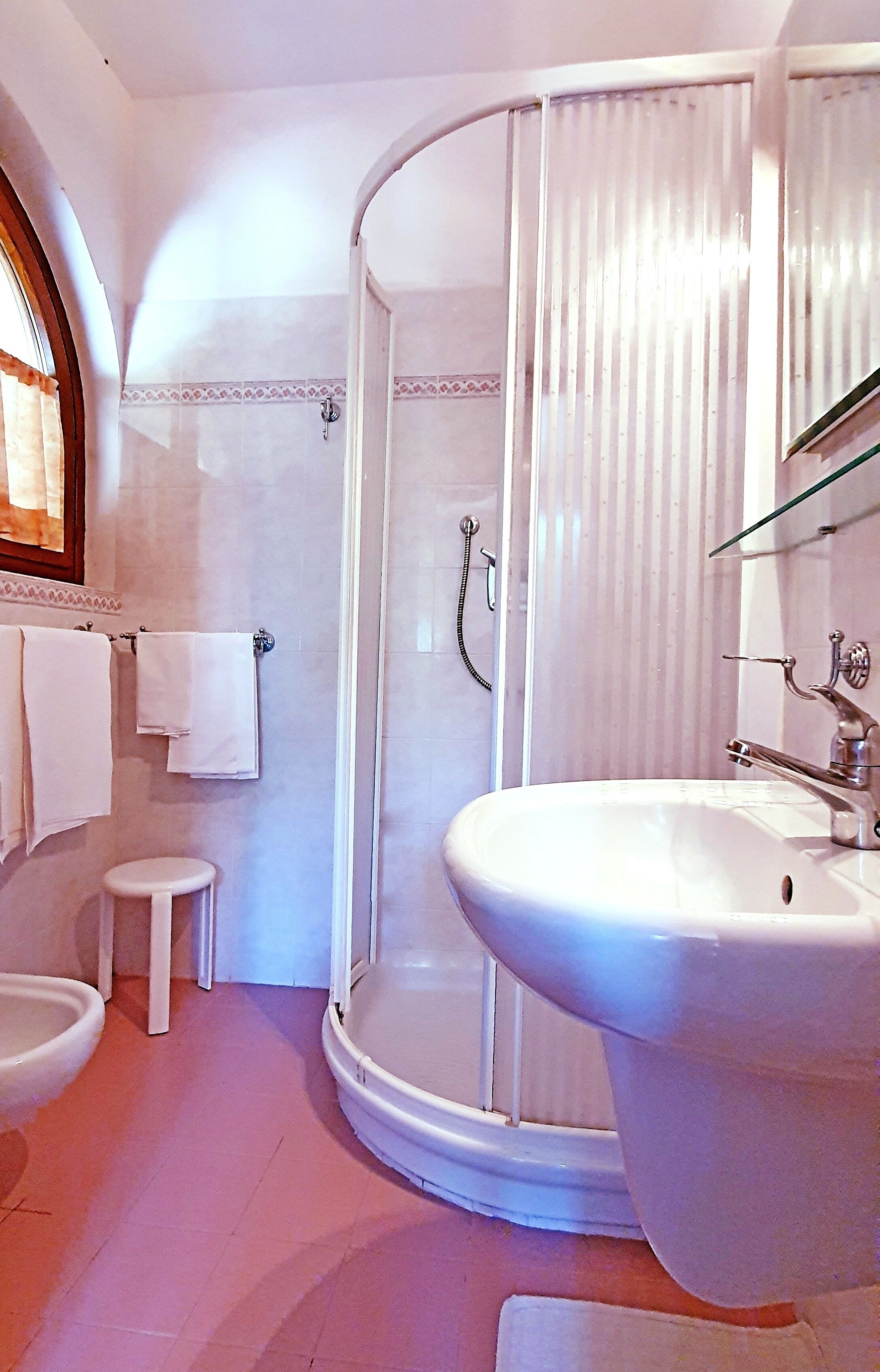 Une salle de bain avec une douche d’angle aux parois incurvées, un lavabo sur colonne et un petit tabouret.