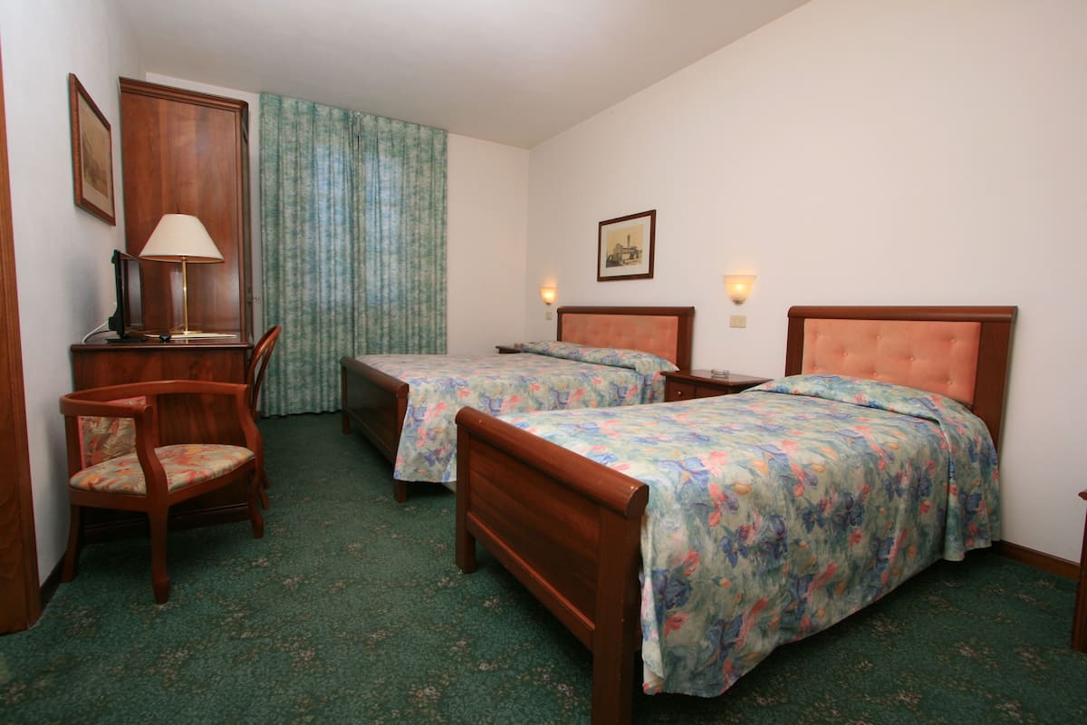 A hotel room with two single beds, a wooden chair, a desk with a lamp, and a picture on the wall.