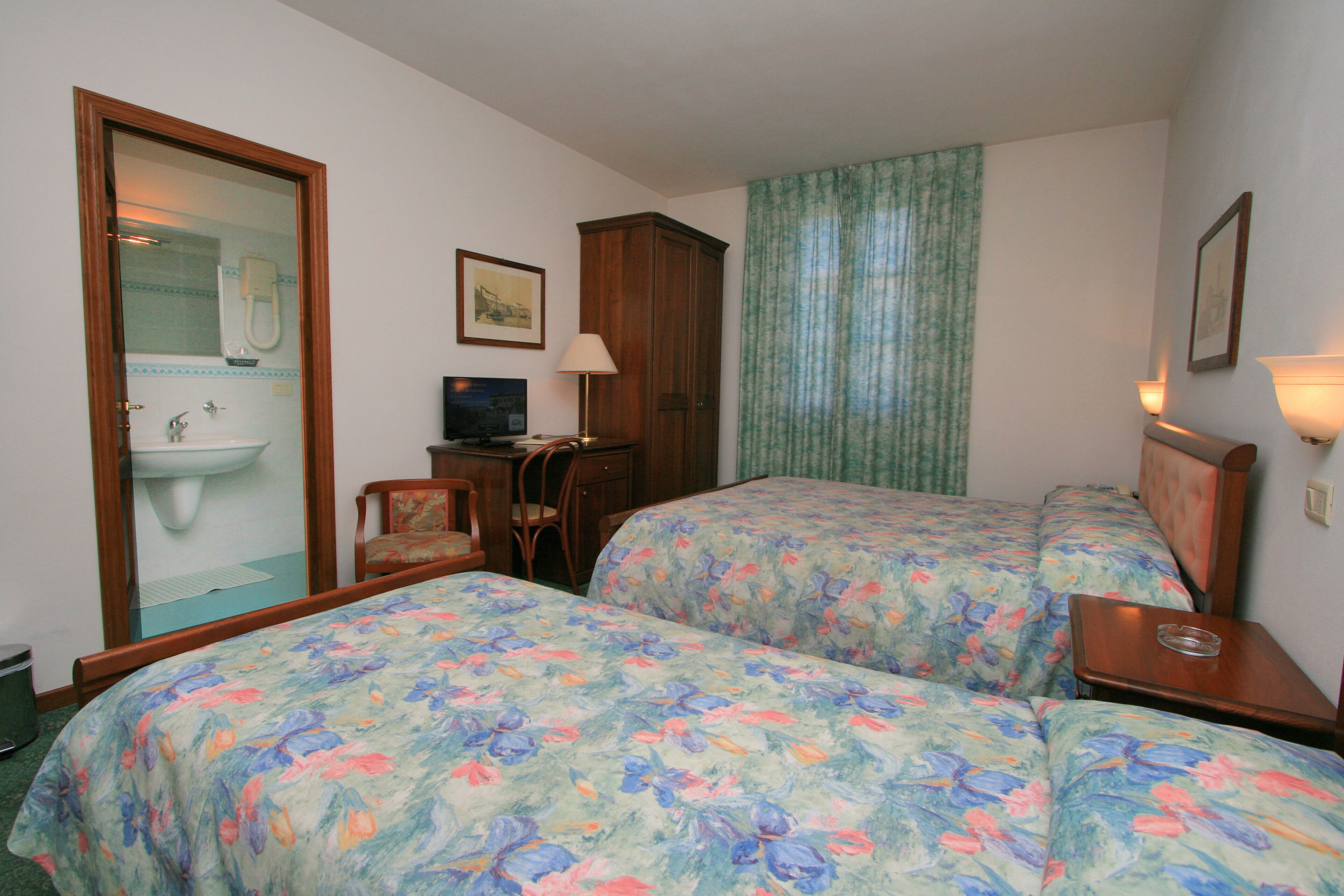 A hotel room with two beds, a desk, a chair, a TV, a cabinet, and a bathroom visible through a glass door.