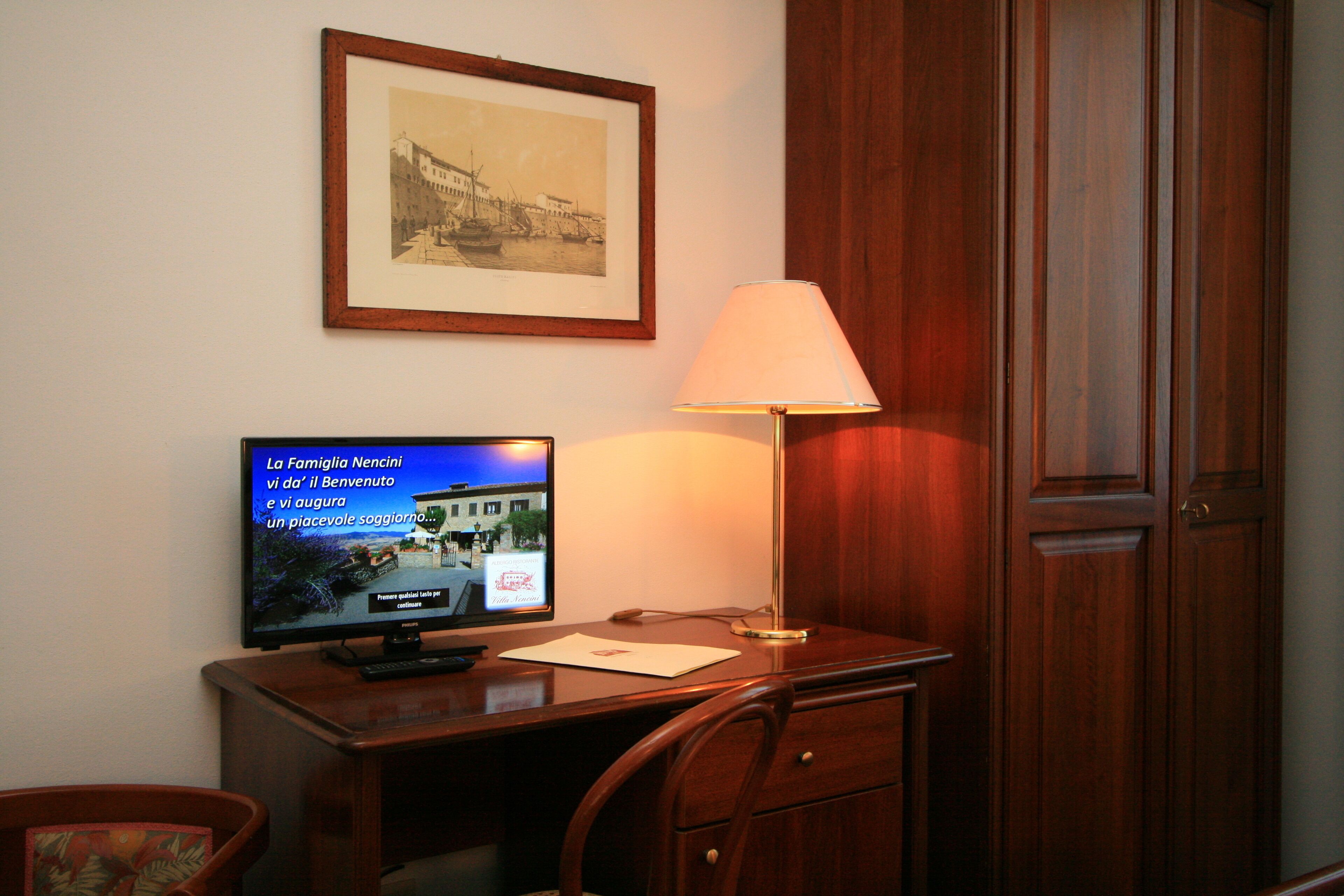 A hotel room with a wooden desk, a television displaying a news channel, a framed picture on the wall, and a wooden wardrobe.