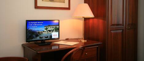 A hotel room with a wooden desk, a television displaying a news channel, a framed picture on the wall, and a wooden wardrobe.