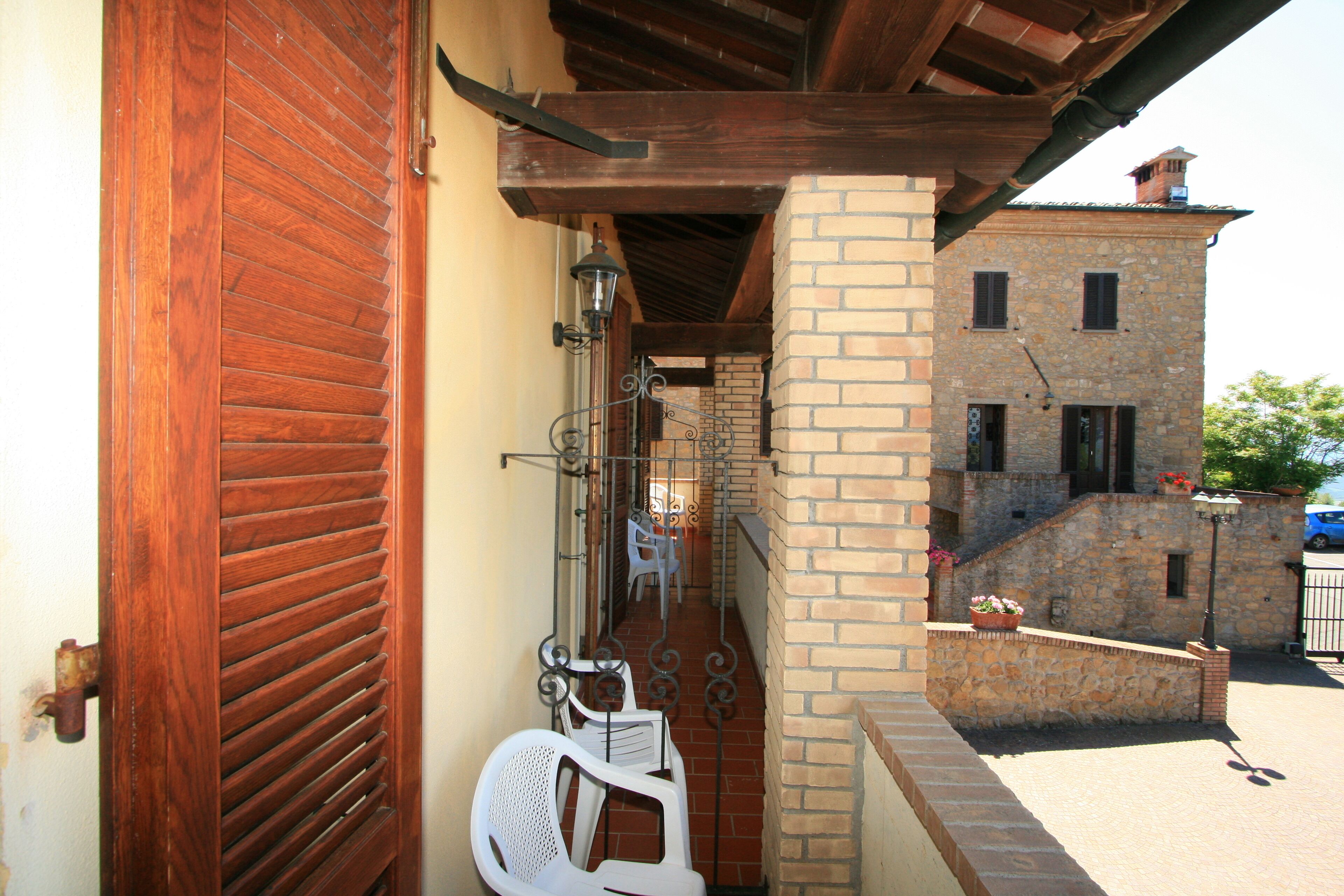 Un estrecho pasillo exterior con una puerta de madera, sillas de plástico blanco y un edificio de piedra al fondo.
