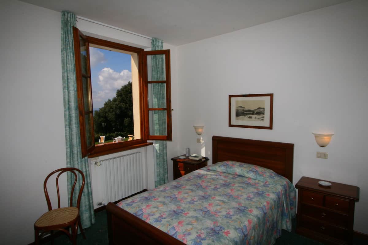 A bedroom with a wooden bed, a chair, a bedside table, a nightstand, a window with curtains, a framed picture on the wall, and a radiator.