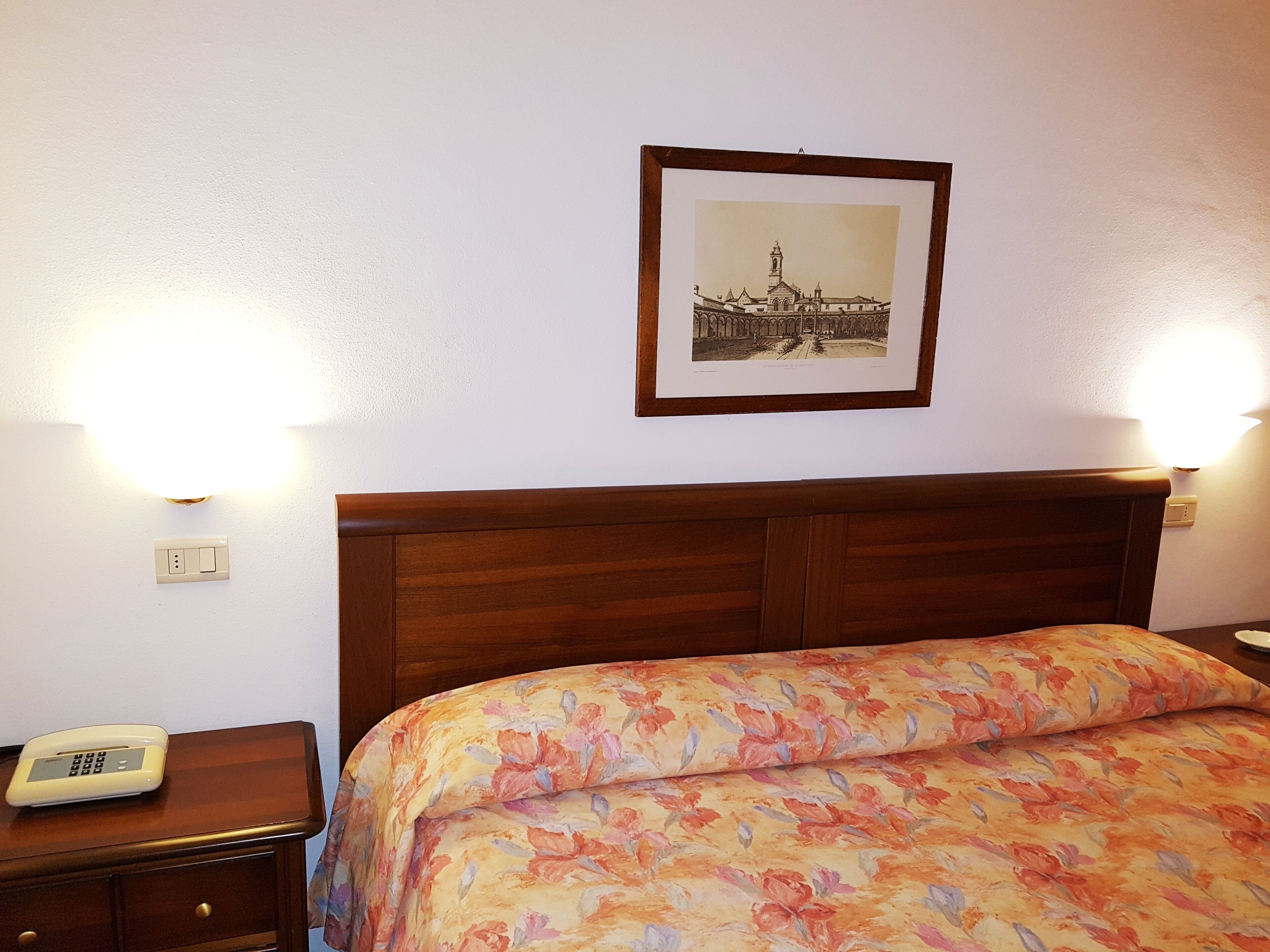 Ein Hotelzimmer mit einem hölzernen Kopfteil, einer blumigen Tagesdecke, einem Nachttisch mit Telefon und einem gerahmten Bild an der Wand.
