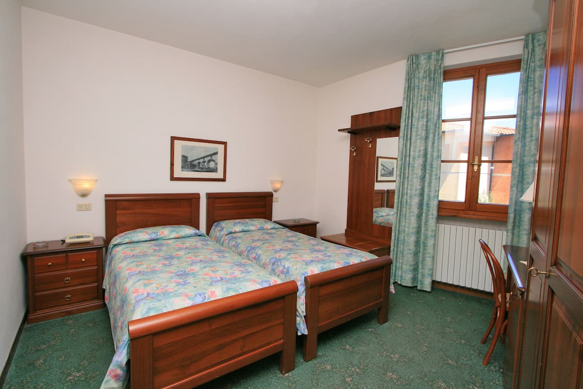 A bedroom with a wooden bed, two bedside tables, a nightstand, a chair, a window with curtains, and a framed picture on the wall.
