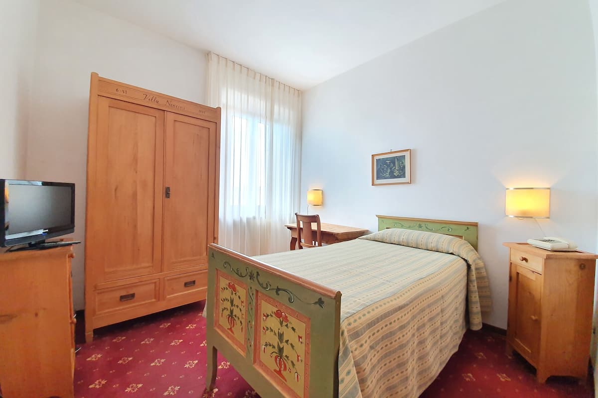 A bedroom with a bed, a wooden wardrobe, a desk with a chair, a television, and a wall-mounted lamp.