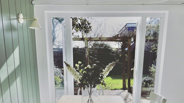 A dining table with chairs, a vase with flowers, and a view of a garden through a glass door. - Island House is a spacious 4 bedroom detached house (West Mersea)