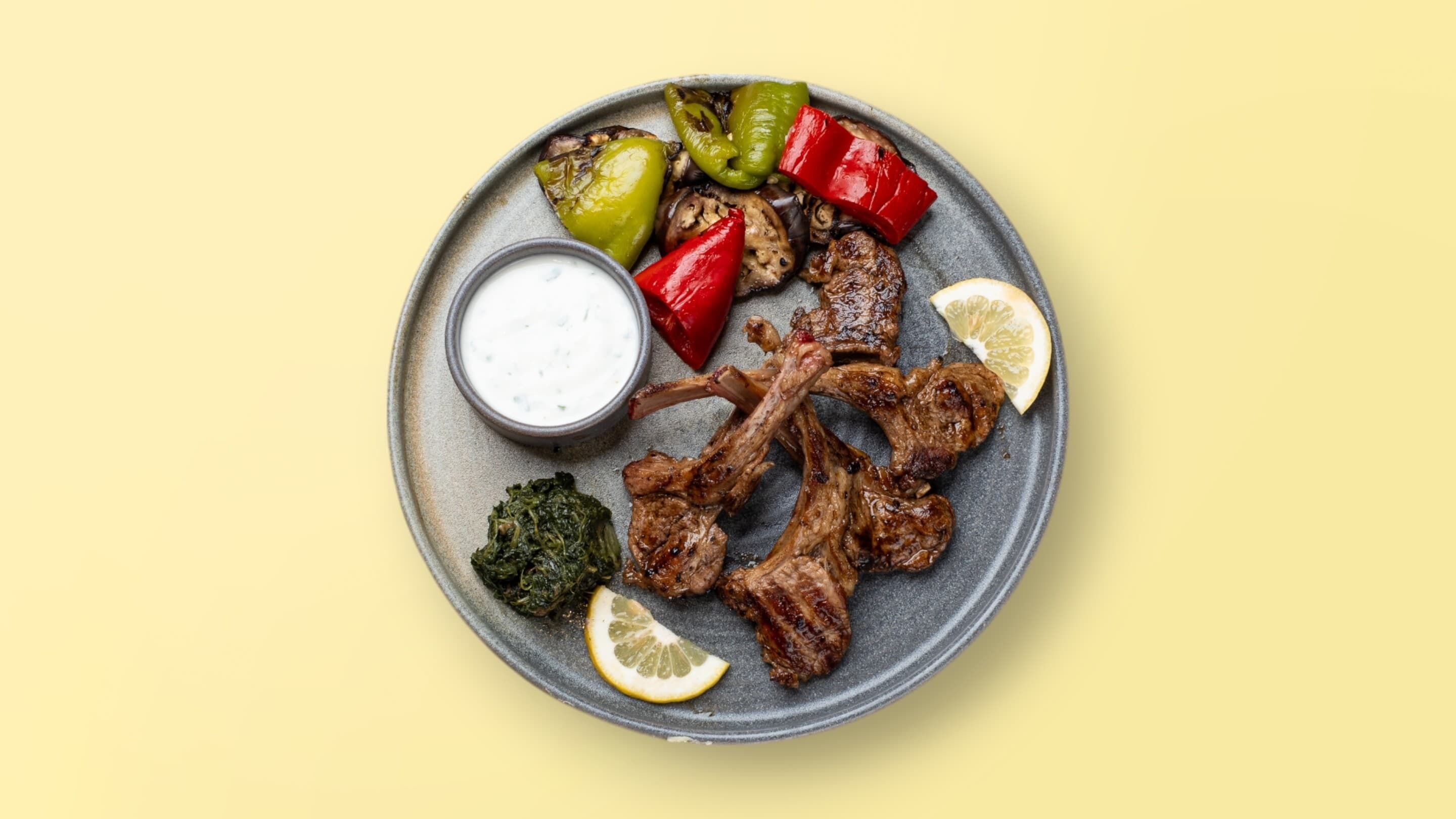 Un plato con chuletas de cordero a la parrilla, verduras asadas, guarnición de vegetales verdes y un bol de salsa.