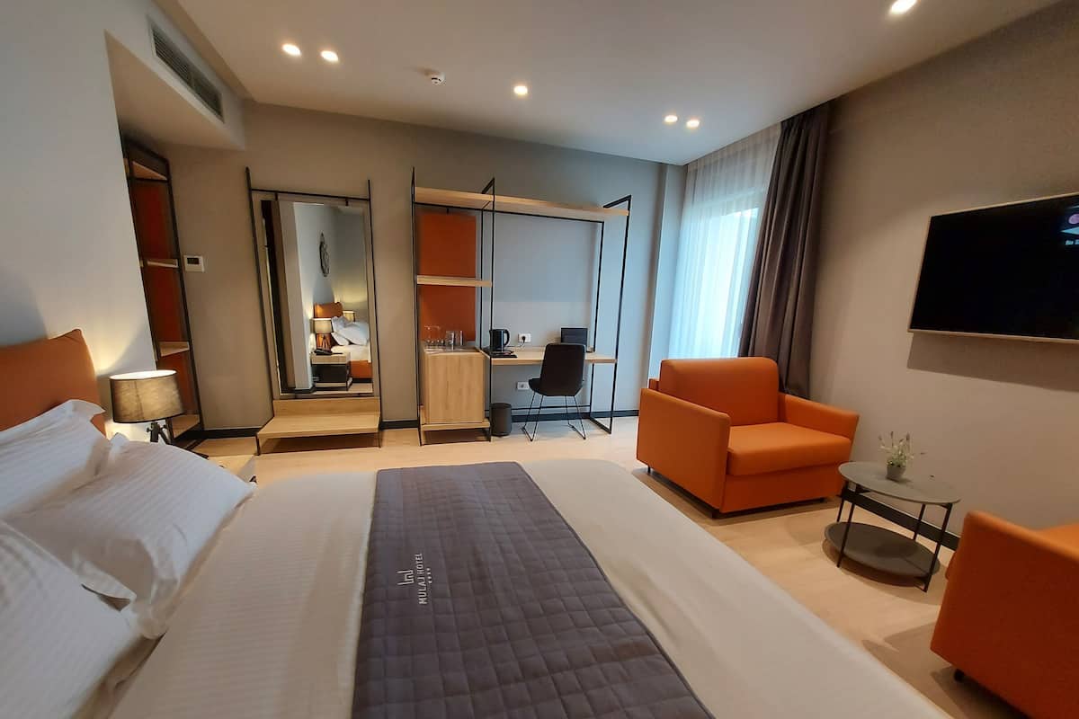 A hotel room with a large bed, an orange armchair, a desk with a chair, a TV mounted on the wall, and a mirror.