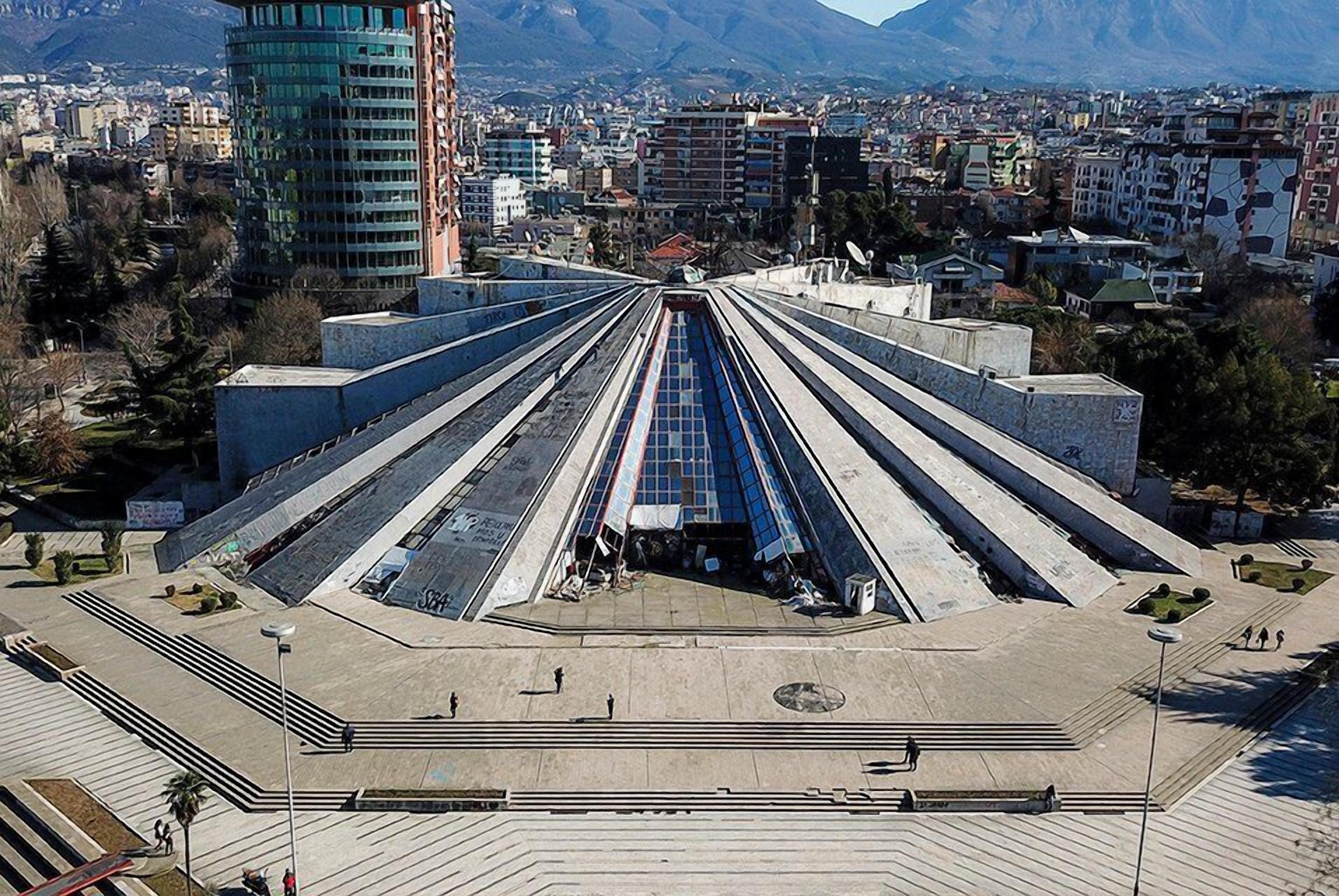 A large public square with a central monument, surrounded by buildings and mountains in the background.