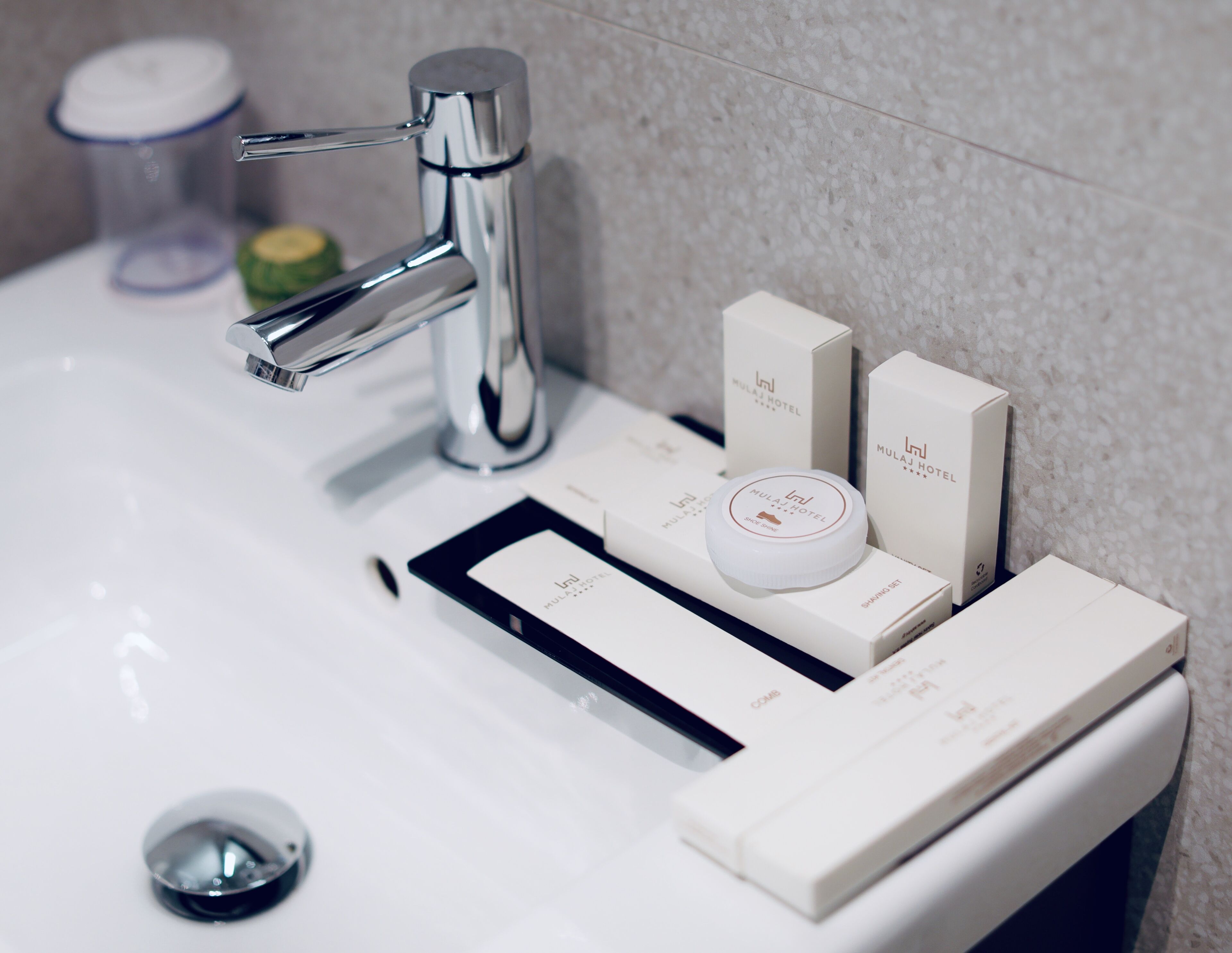 A bathroom sink with a chrome faucet, a soap dispenser, and toiletries including a box labeled "Mulaj Hotel."