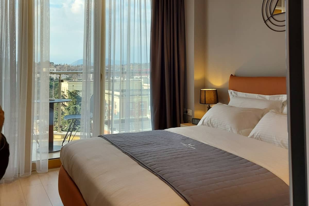 A hotel room with a large bed, a view of the city, and a balcony with a table and chairs.