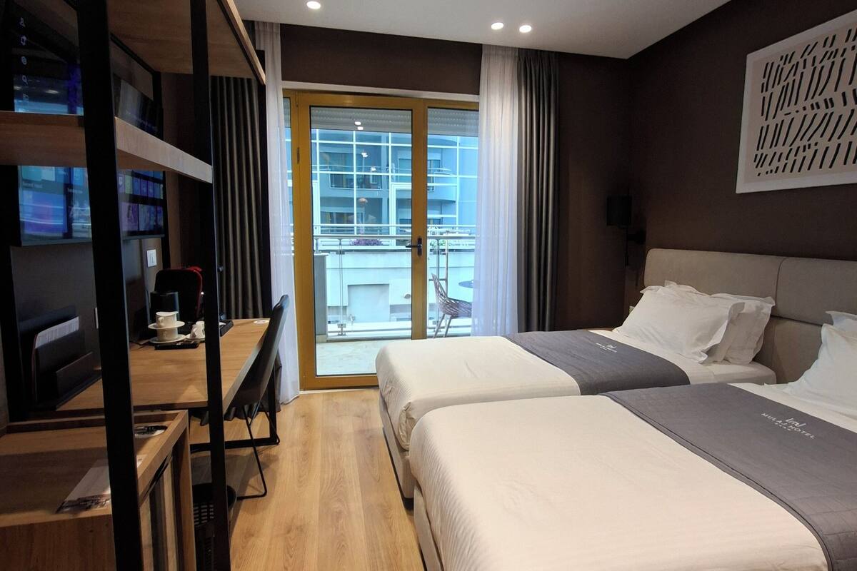 A modern hotel room with two beds, a wooden desk, and a view of the city through large windows.