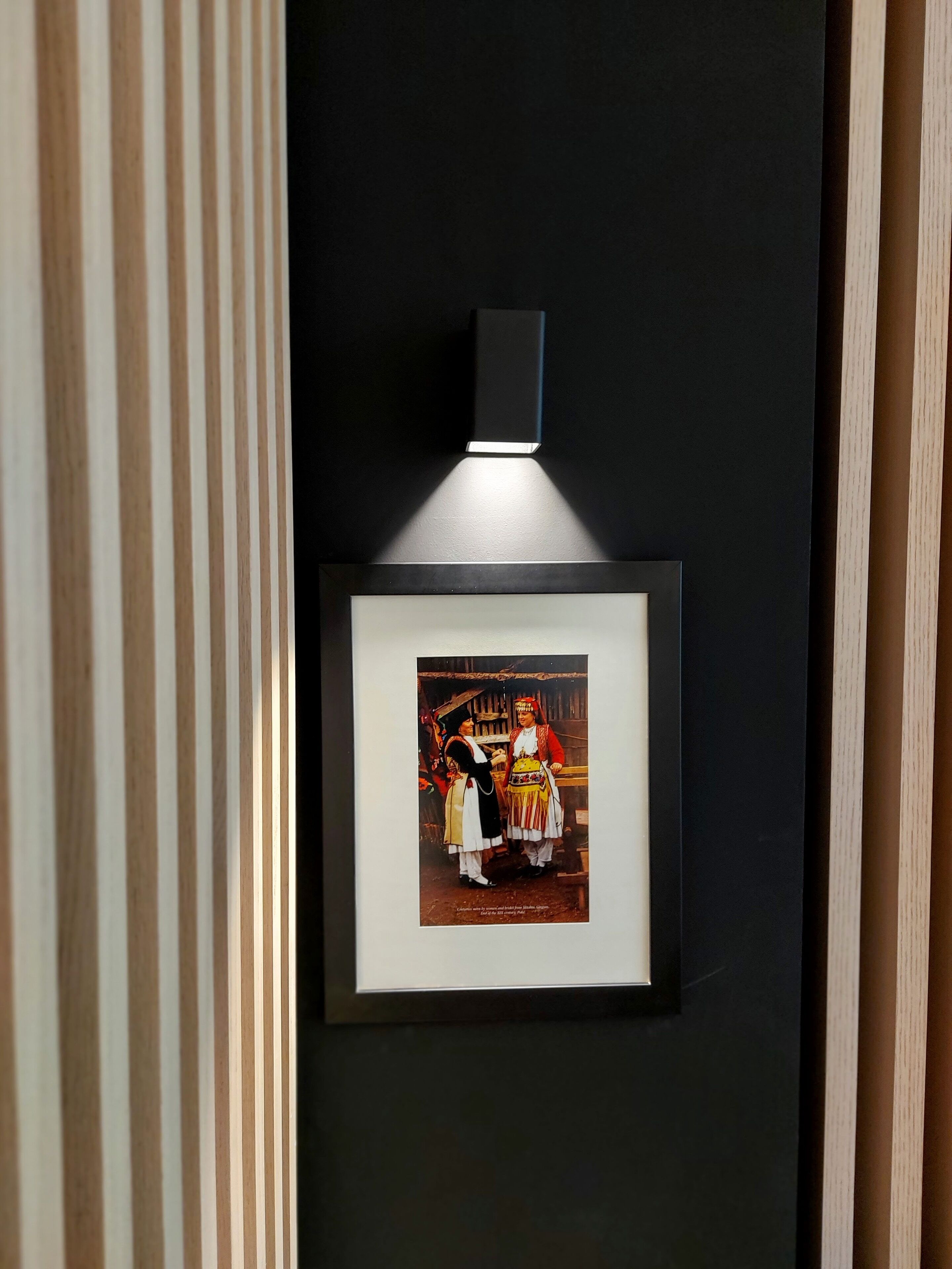 Een ingelijste foto van twee personen in traditionele kledij, bevestigd aan een donkere muur met een lichtbron erboven.
