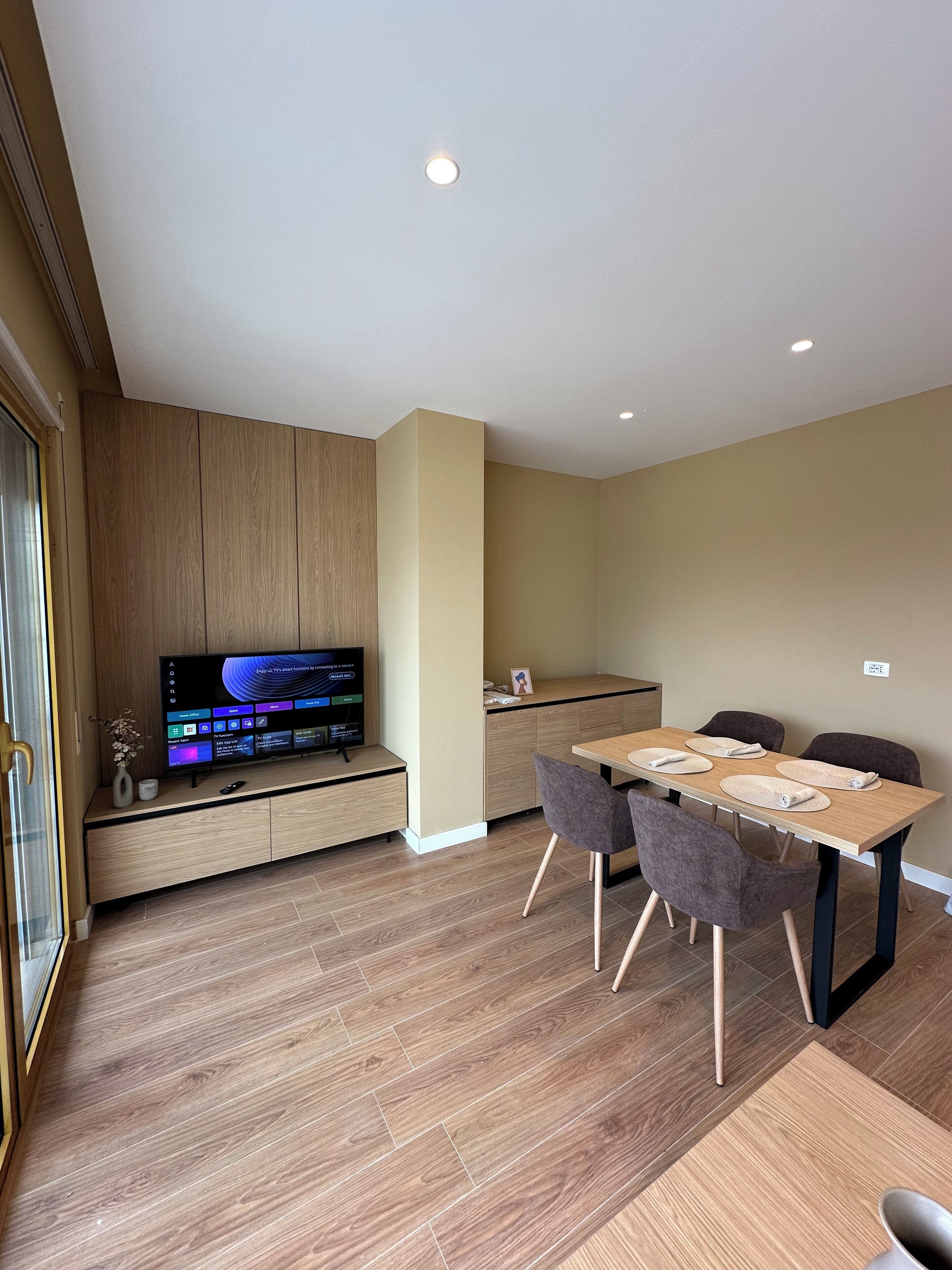 A modern living room with a flat-screen TV, a dining table with four chairs, and a kitchen area with wooden cabinets.